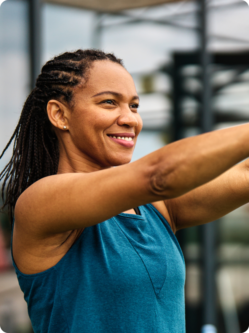 Smiling woman in a teal vest stretching her arms out during an outdoor workout