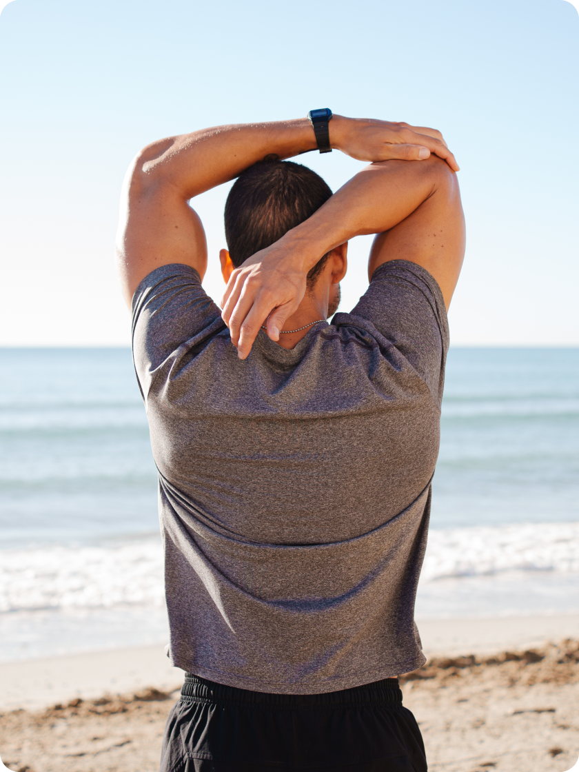 Man in a grey T-shirt stretching his arms behind his head on a sunny beach