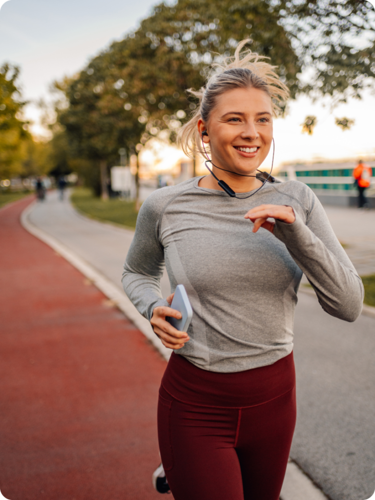 Smiling woman jogging along a park path, wearing earphones and holding her phone