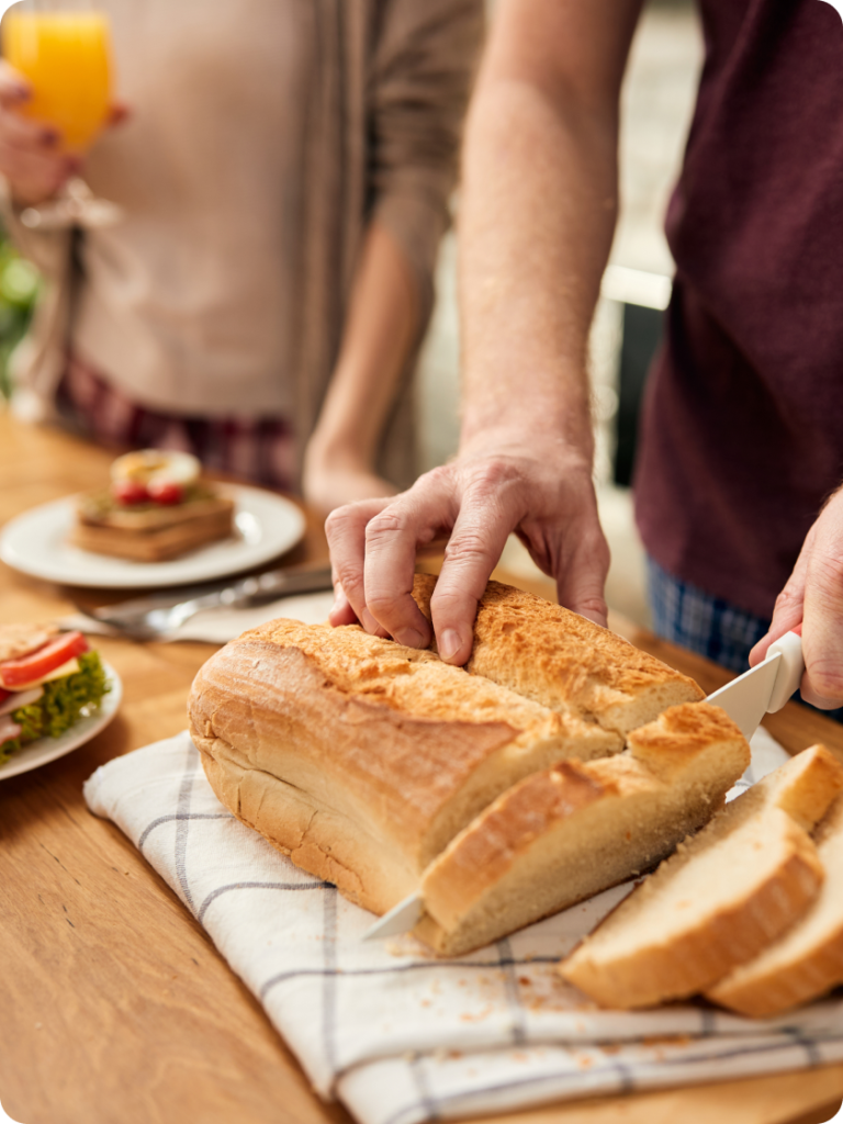 Man slicing a loaf of fresh bread in a kitchen, with salad plates and a glass of orange juice nearby